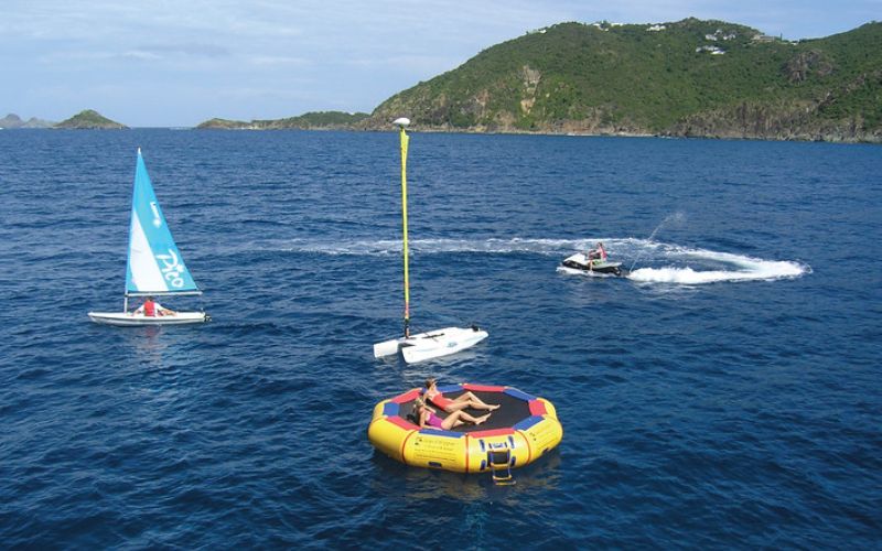 This image captures a lively water sports scene offered by SeaDream Yacht Club, featuring guests enjoying activities in a secluded bay. A person relaxes on a floating inflatable platform, while others sail a small boat and ride a jet ski in the vibrant blue water. The lush green hills in the background provide a serene natural setting for these aquatic adventures.
