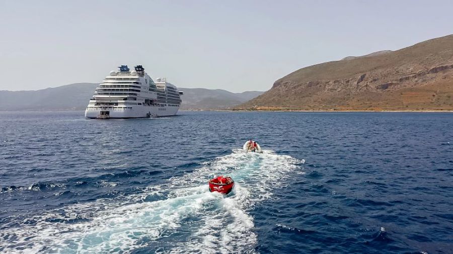 During Seabourn cruise's Marina Day, the ship is anchored near a rugged coastline, with guests enjoying water sports activities such as riding in red speedboats, creating a sense of adventure against the tranquil blue sea.