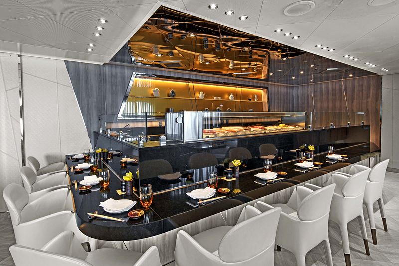 The sleek and modern Sushi restaurant aboard Seabourn Ovation, with its striking black and gold sushi bar, white contemporary chairs, and sophisticated dining ambiance, invites guests to a luxurious sushi experience at sea.