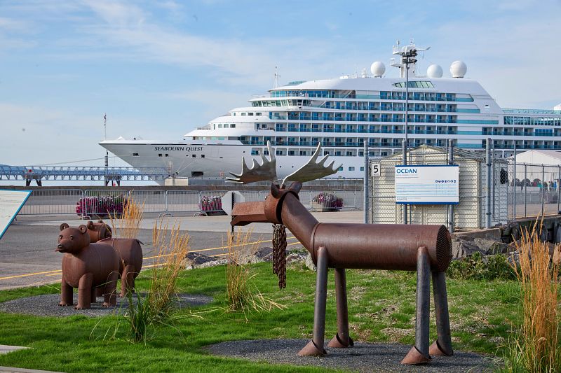 The Seabourn Quest cruise ship docked at a Canadian port, with playful metal sculptures of a bear and a moose in the foreground, blending the marvel of engineering with local art and wildlife themes.