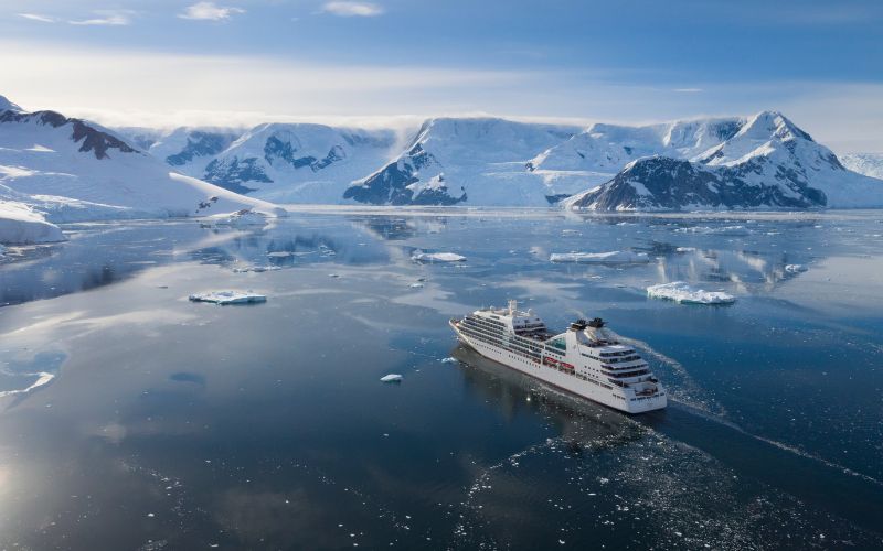 An aerial shot captures the Seabourn Quest cruise ship navigating through the icy waters of Neko Harbor, Antarctica. Surrounded by scattered ice floes and embraced by snow-covered mountains, the ship's journey highlights the pristine and remote beauty of the Antarctic landscape.