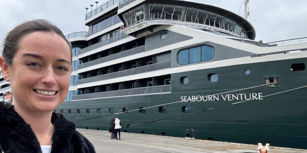 Cruise Mummy with a warm smile, wearing a black jacket, takes a selfie with the large, green-hulled Seabourn Venture cruise ship in the background. The ship's multiple decks are visible, along with its name clearly printed on the side. In the foreground, a few people can be seen walking near the docked ship under a cloudy sky.