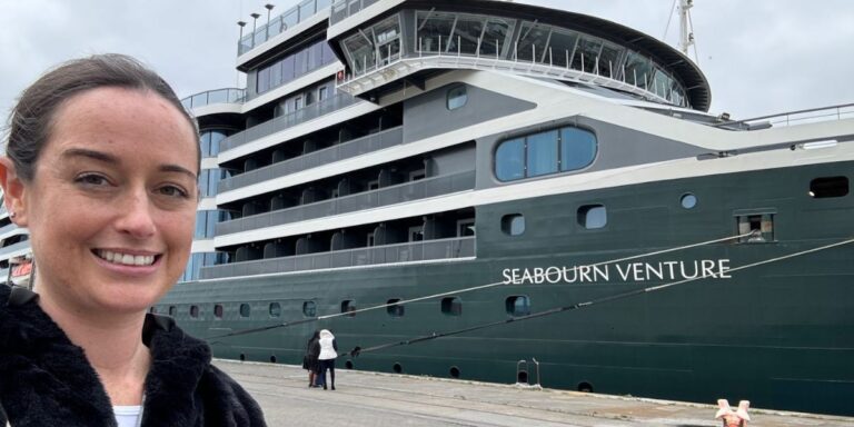 Cruise Mummy with a warm smile, wearing a black jacket, takes a selfie with the large, green-hulled Seabourn Venture cruise ship in the background. The ship's multiple decks are visible, along with its name clearly printed on the side. In the foreground, a few people can be seen walking near the docked ship under a cloudy sky.