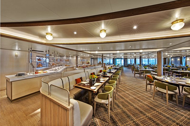 The Colonnade dining area on Seabourn Venture, with a buffet spread in the foreground, offers a warm, inviting space featuring plush seating, elegant table settings, and natural light streaming through large windows, embodying sophisticated comfort.