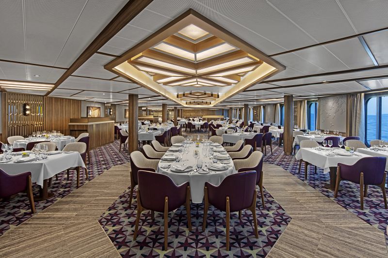 The Restaurant on the Seabourn Venture cruise ship presents an elegant dining space with geometric carpet designs, deep purple chairs, white tablecloths, and an impressive geometric ceiling light feature, all set against ocean views through the large windows.