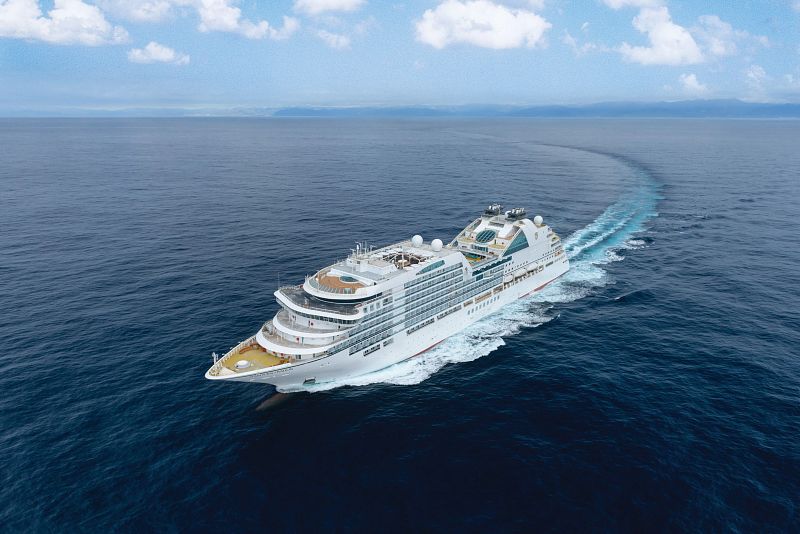 The Seabourn Ovation luxury cruise ship cuts through the deep blue sea, leaving a frothy wake behind, with its white hull and distinctive yellow accents under a vast, open sky.