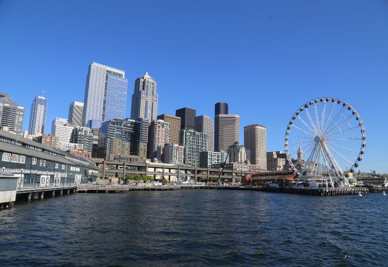 A vibrant view from a cruise ship of downtown Seattle's skyline, punctuated by the iconic Great Wheel Ferris wheel on the pier, set against a clear blue sky, showcasing the city's blend of urban and recreational spaces.