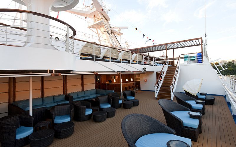 The Serenity Deck on the Carnival Dream cruise ship during the day, featuring cozy blue-cushioned rattan loungers, wooden flooring, white railings, and festive flags fluttering above, offering a peaceful outdoor lounging experience.