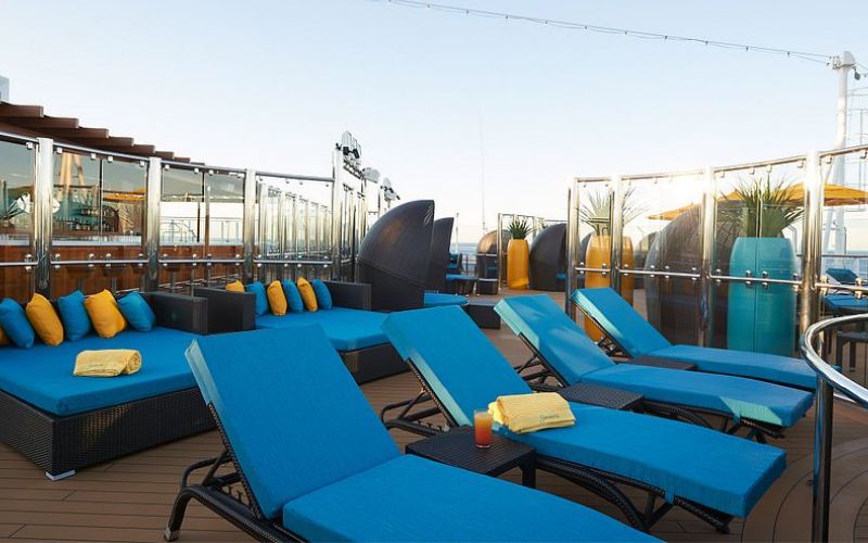 Inviting Serenity deck on the Carnival Panorama featuring vibrant blue sun loungers with plush yellow towels, contemporary wicker sofas, and transparent windbreaks, all set for relaxation and offering panoramic views at sea.