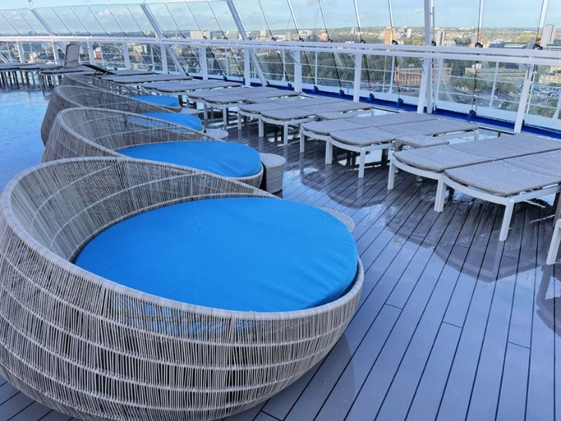 Relaxing Serenity deck on a Carnival cruise ship featuring modern circular loungers with vibrant blue cushions, sleek sunbeds, and a translucent dome overhead, all with a city skyline visible in the distance.