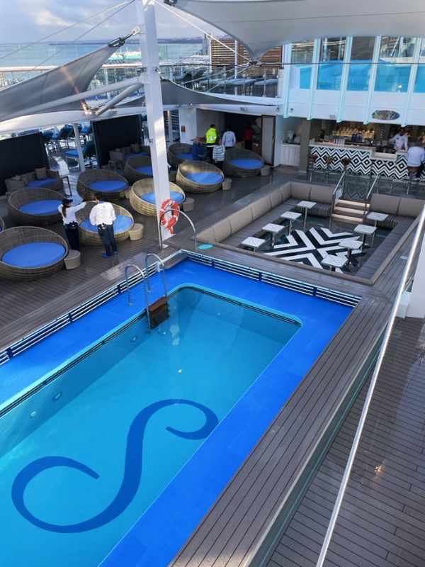 Elevated view of the Serenity deck on the Carnival Celebration cruise ship, featuring the iconic Serenity logo in the pool, with plush round loungers, a poolside bar, and a lounge area with modern geometric designs, all set for a relaxing escape at sea.