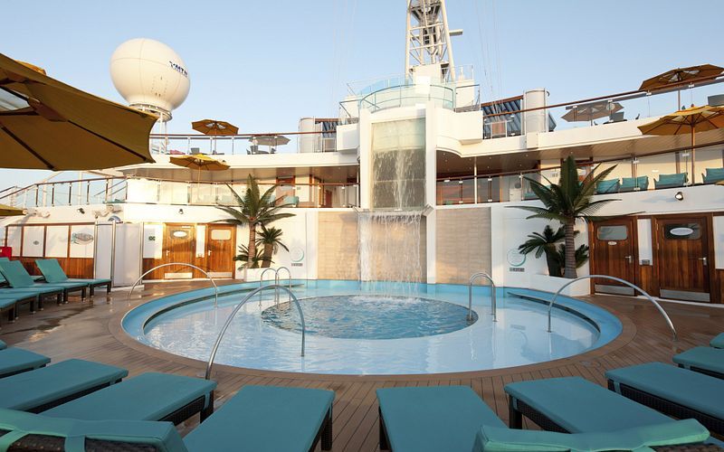 Tranquil Serenity pool area on the Carnival Sunshine with a cascading water feature, turquoise sunbeds, lush palm trees, and umbrellas, flanked by the ship's white superstructure and a clear blue sky overhead.