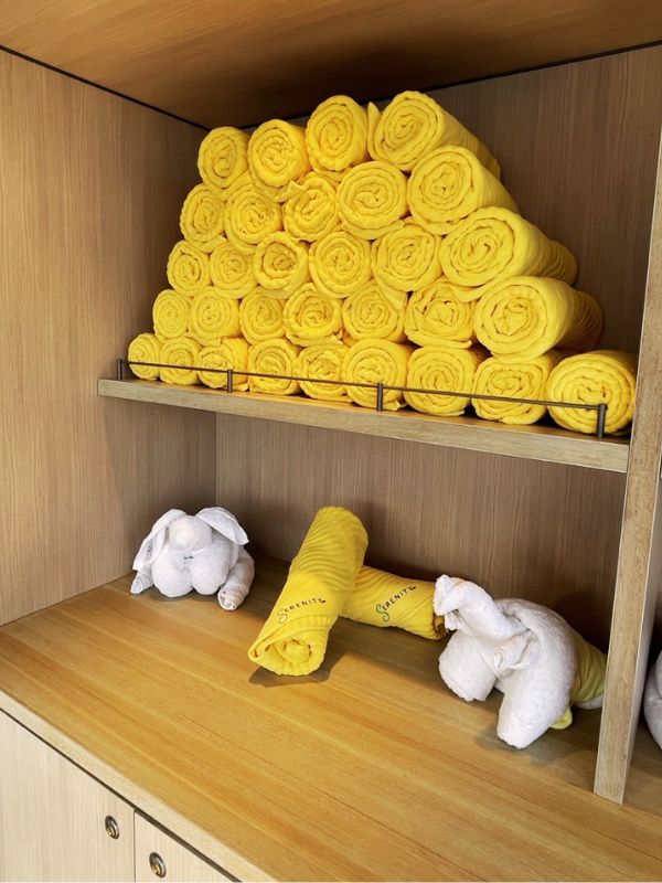 A cozy display of yellow 'Serenity' branded towels rolled into floral shapes, neatly stacked on a wooden shelf beside playful white towel animals, creating a charming and welcoming atmosphere in the Serenity area on the Carnival Celebration.