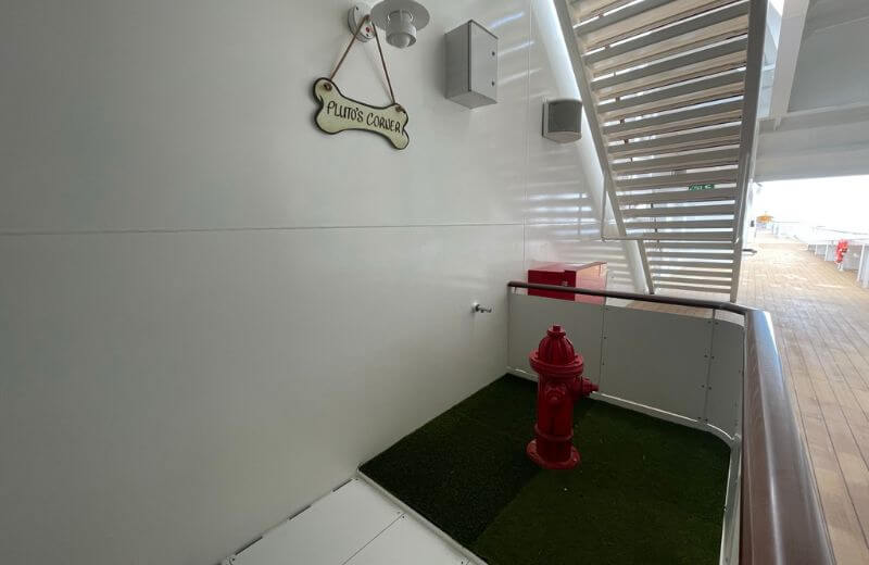 "Pluto's Corner," a designated relief area for service dogs on the Disney Wish cruise ship, featuring synthetic grass and a bright red fire hydrant. The clean, white walls and modern design elements, combined with the practical outdoor setting, reflect a thoughtful consideration for the comfort of service animals on board.