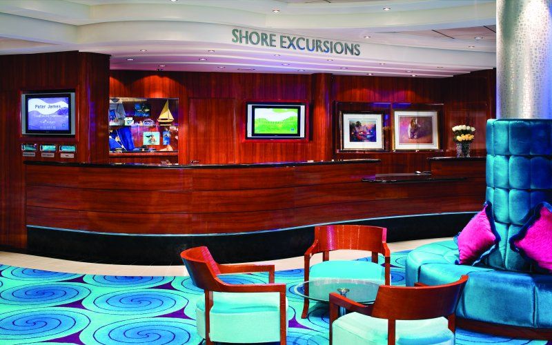 The Shore Excursions desk on a Norwegian Cruise Line ship, featuring a polished wood counter, informational screens, and plush seating in a cozy lounge area with vibrant blue carpeting, ready to assist guests with their onshore adventure planning.