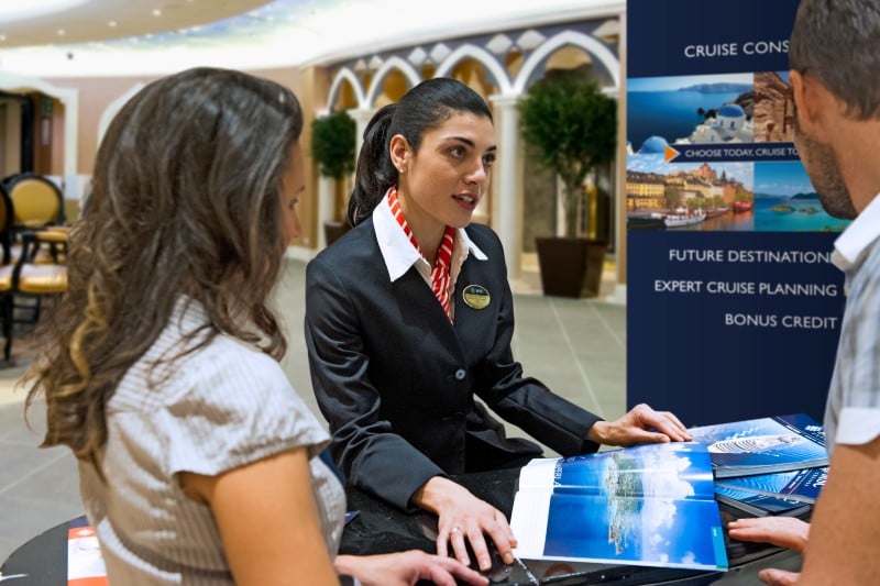 A shore excursion manager is discussing shore excursions with a couple at a travel desk, with brochures and promotional materials displayed. The setting appears to be an upscale, elegant lobby, emphasizing personalized service and luxury travel planning.