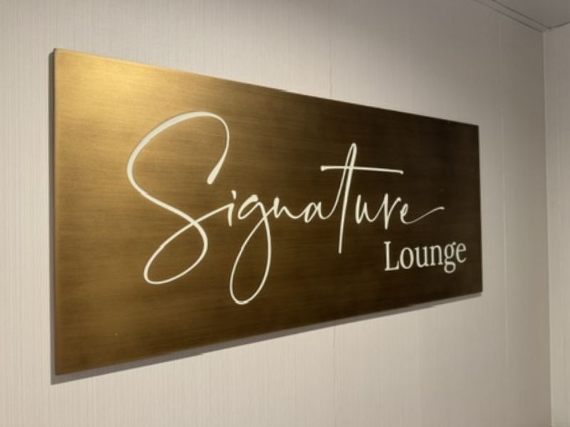 The image shows a sign for the "Signature Lounge" on Princess Cruises' Sun Princess. The sign is mounted on a wall and features elegant, cursive white lettering on a sleek, bronze-colored background, indicating the entrance to this exclusive lounge area aboard the ship. The design of the sign reflects a sense of sophistication and luxury, likely mirroring the ambiance of the lounge itself.