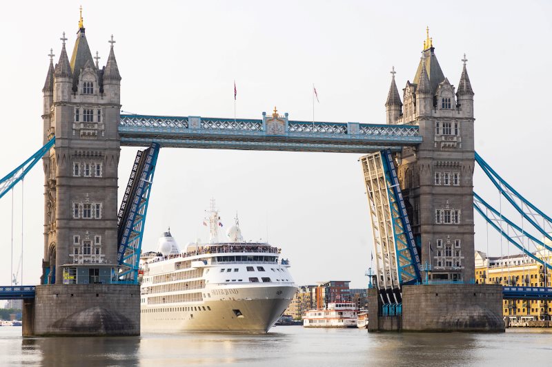 The Silver Spirit cruise ship from Silversea Cruises passing under the iconic Tower Bridge in London, which is raised to allow the vessel through, highlighting the grandeur of both the ship and the landmark bridge.