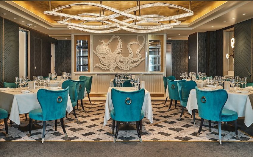 The Il Terrazzino restaurant on board the Silver Endeavour features a luxurious interior with teal velvet chairs, geometric-patterned flooring, grand chandeliers, and a striking white octopus wall art, creating a lavish dining ambiance.