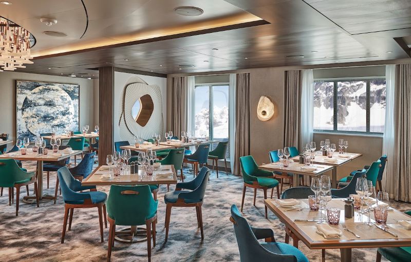 The Restaurant on the Silver Endeavour features a sophisticated interior with teal and beige chairs, elegant table settings, contemporary art pieces, and panoramic windows offering a glimpse of the snowy landscape outside, providing guests with a refined dining experience at sea.