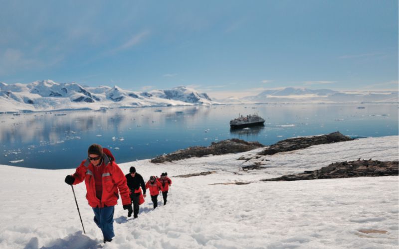 This image captures Silversea guests hiking through a pristine, snowy landscape in Antarctica, wearing bright red parkas for warmth and visibility. In the background, an expedition cruise ship floats on calm, icy waters surrounded by dramatic snow-covered mountains under a clear blue sky. The scene showcases an adventurous and serene exploration of this remote polar region.
