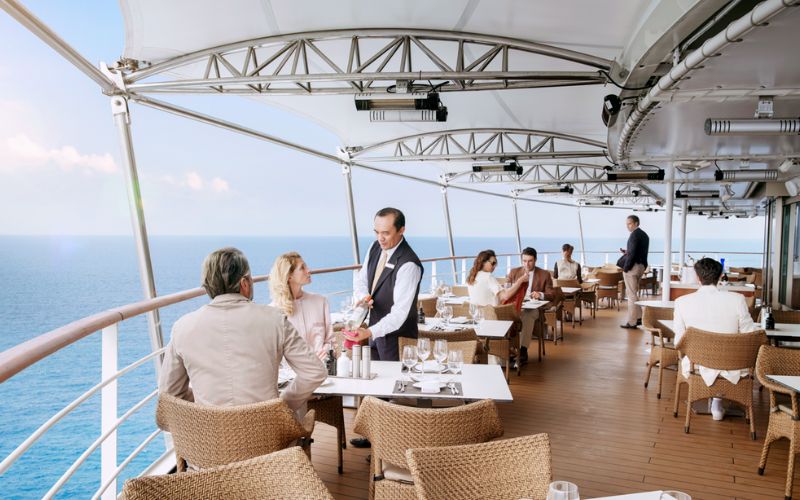 This image showcases an elegant outdoor terrace restaurant on a Silversea cruise ship, with a stunning view of the ocean. Guests dine at tables set with crisp white linens while attentive waitstaff provide personalized service. The airy, open design and serene blue horizon create a luxurious and tranquil dining experience.