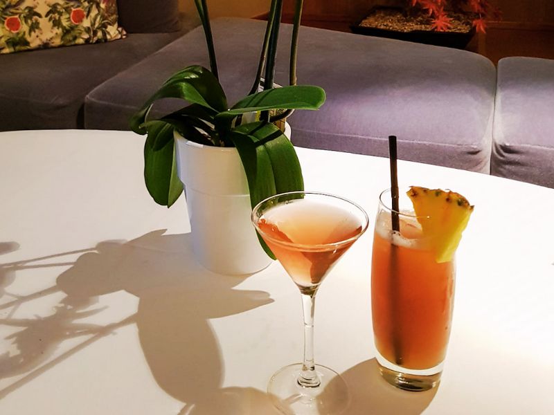 A Singapore Sling cocktail on a cruise ship, served in a tall glass garnished with a pineapple slice and accompanied by a martini glass with a pink drink. The drinks are placed on a white table next to a potted plant, with a cozy seating area in the background, creating a sophisticated and relaxed ambiance.