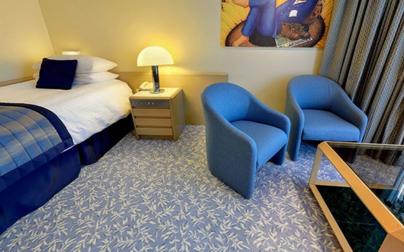 A Single Balcony Suite on the Fred Olsen Balmoral cruise ship, featuring a cozy bed with white linens and blue accents. The room includes a small sitting area with two blue armchairs and a glass-top coffee table, as well as a nightstand with a lamp and a framed artwork on the wall. The decor combines soft, inviting tones with a comfortable layout, perfect for relaxation.