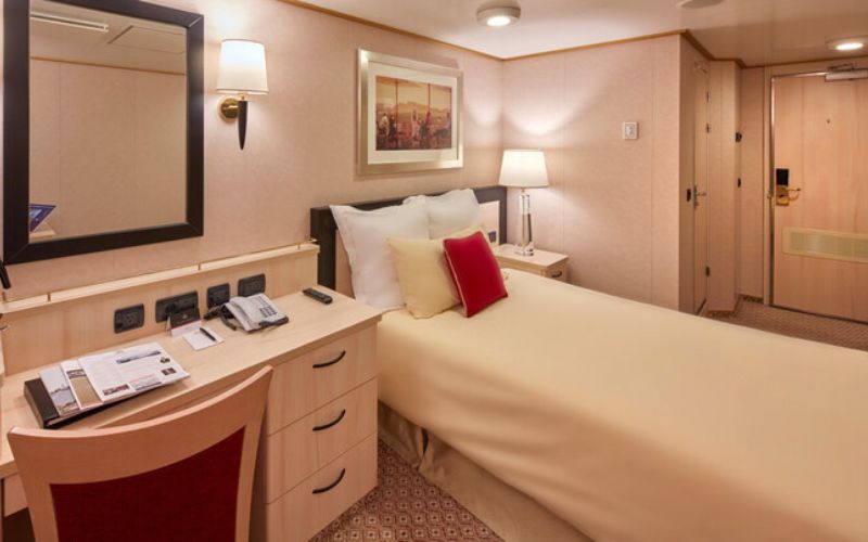 A cozy Single Inside stateroom on board the Queen Elizabeth cruise ship, featuring a neatly made bed with white and red accent pillows. The room includes a desk with a mirror, a telephone, and reading materials, as well as a nightstand with a lamp, creating a warm and inviting ambiance with elegant decor.