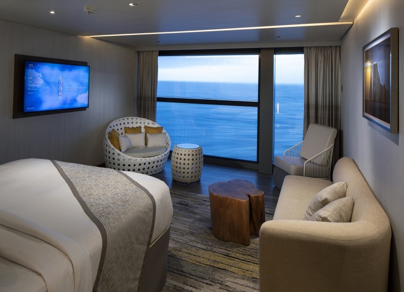 Cozy Sky Suite with Infinite Veranda on the Celebrity Flora, featuring a plush bed, a comfortable seating area with chic furnishings, a widescreen TV, and floor-to-ceiling sliding glass doors that open up to uninterrupted views of the ocean.