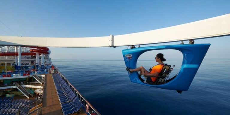 A passenger enjoys the SkyRide attraction on Carnival Panorama, pedaling a hanging bike along a track high above the ship's deck, offering expansive ocean views and a fun, aerial cycling adventure at sea.