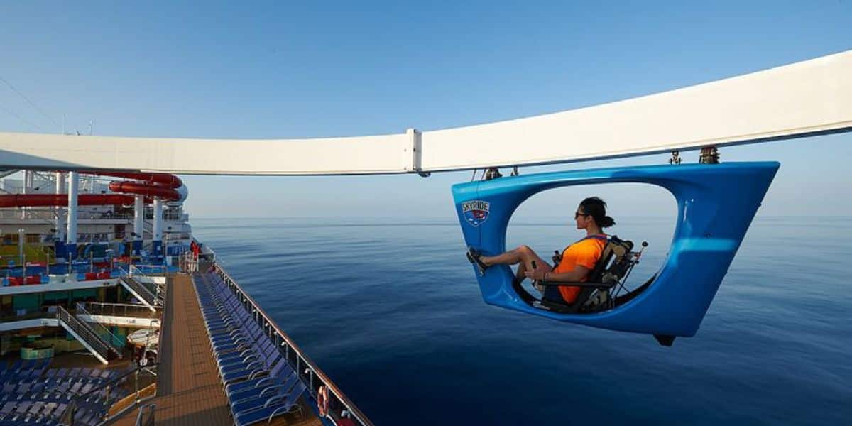 A passenger enjoys the SkyRide attraction on Carnival Panorama, pedaling a hanging bike along a track high above the ship's deck, offering expansive ocean views and a fun, aerial cycling adventure at sea.