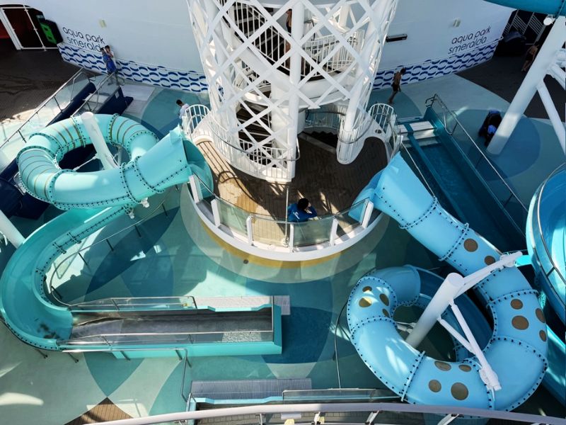 Overhead view of the intricate water slide complex on Costa Smeralda, featuring a series of intertwined slides in shades of blue, with vacationers enjoying the sun-drenched deck area below.