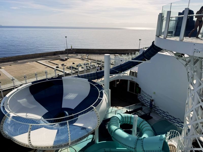 View from the top of a spiraling water slide on the Costa Smeralda cruise ship, overlooking the deck and the calm blue sea beyond, with passengers preparing to enjoy the thrill of the slide.