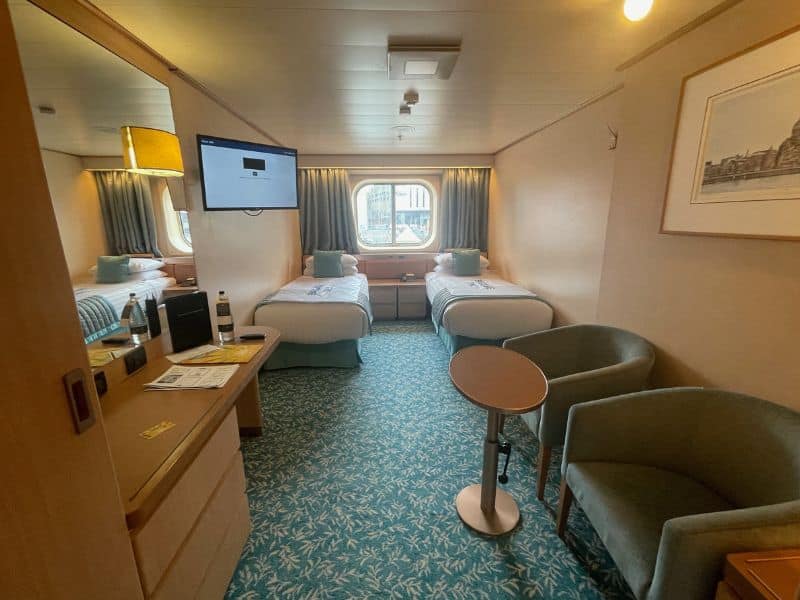 A comfortable solo cabin on a Fred. Olsen cruise ship, featuring two twin beds with light blue accents and a large window offering an outside view. The room includes a small sitting area with two chairs and a round table, a desk with a lamp and a flat-screen TV mounted on the wall, creating a cozy and inviting atmosphere.