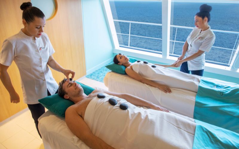 A couple indulges in a relaxing spa treatment at Carnival's Cloud 9 Spa, with therapists gently performing a head massage and a hand treatment. They are lying on massage tables with ocean views, experiencing a serene escape with the endless sea as their backdrop.