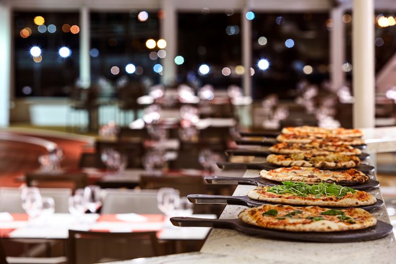 The Spaccanapoli pizzeria on a Silversea cruise ship, captured in the evening with a lineup of freshly baked pizzas ready to be served, evoking a cozy, inviting dining atmosphere with a view of the city lights through the windows.