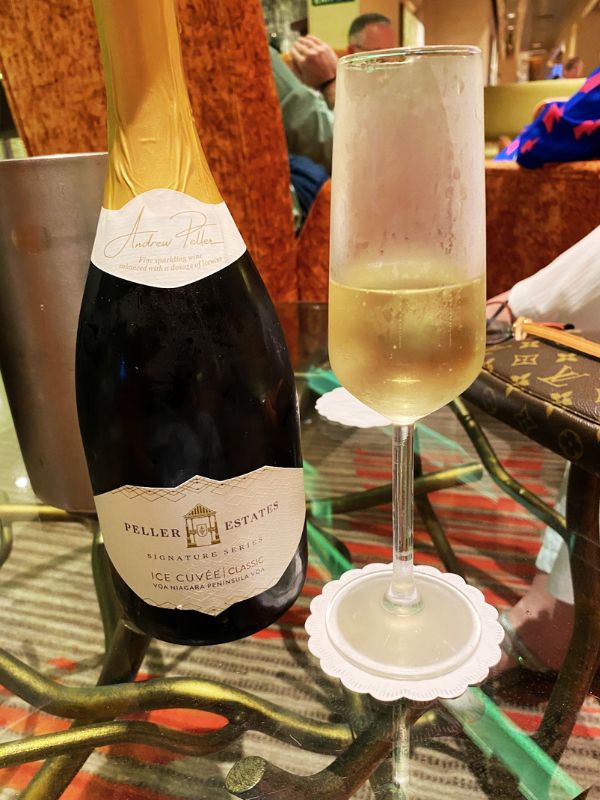 A bottle of Peller Estates Ice Cuvée Classic sparkling wine with a glass filled and placed on a glass table aboard a cruise ship. The setting includes elegant decor with plush seating in the background, creating a refined and relaxed ambiance for enjoying a luxurious drink.