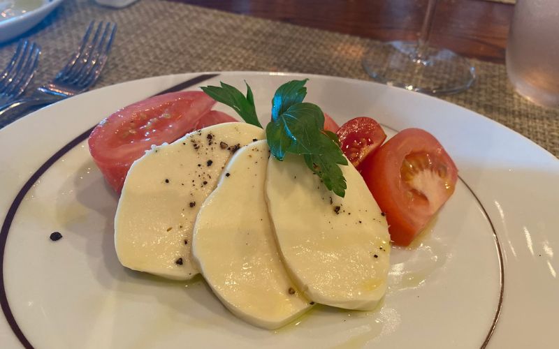 A classic caprese salad with ripe tomatoes, sliced fresh mozzarella, and a sprig of basil, seasoned with black pepper and olive oil, presented on a white plate as part of a specialty dining experience on a Norwegian Cruise Line voyage.