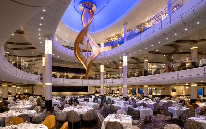 The main dining room on Spectrum of the Seas, featuring elegant, modern decor with two levels of seating. The room is adorned with a striking, twisted sculpture hanging from the ceiling, and tables are set with white tablecloths and place settings, ready for dining guests. Soft, ambient lighting creates a warm and inviting atmosphere.
