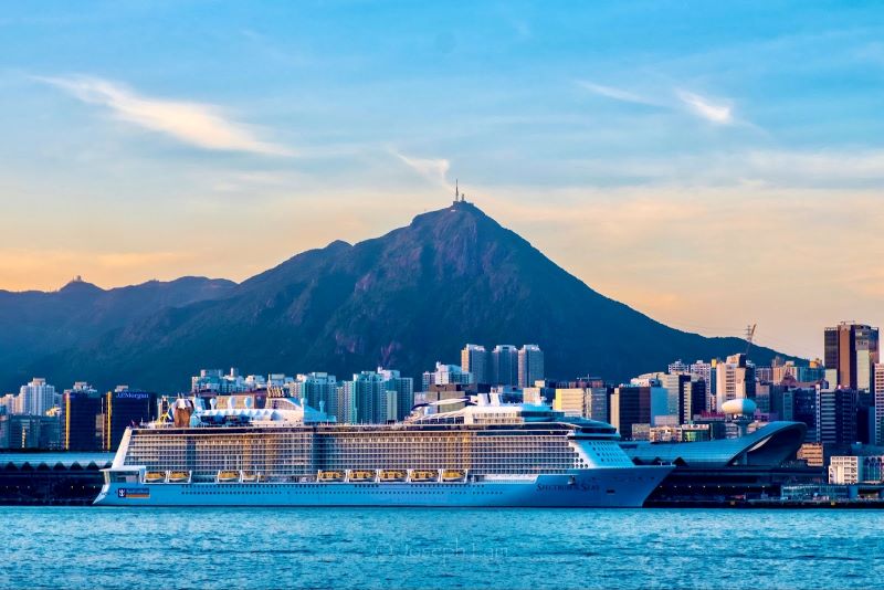 The Spectrum of the Seas cruise ship by Royal Caribbean docked in a harbor at dusk, with the backdrop of a vibrant cityscape and a mountain, under a soft sky transitioning to evening.