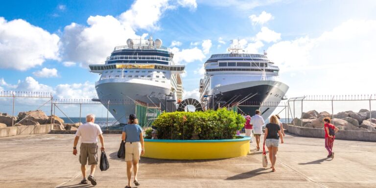 The bustling atmosphere at Port Zante in St. Kitts, with two large cruise ships moored at the terminal. The perspective shows passengers walking along the dock, enjoying the sunny Caribbean weather as they possibly prepare to explore the island or return to the ships after a day out. The ships are impressive in scale, dominating the background, while the foreground features people of various ages in casual vacation attire, emphasizing the tourist-friendly environment. The inclusion of vibrant greenery in a large circular planter adds a touch of the island’s lush natural beauty, contrasting nicely with the industrial elements of the port. This scene effectively portrays Port Zante as a key entry point for visitors to St. Kitts, serving as a gateway to the island's rich culture and scenic landscapes.