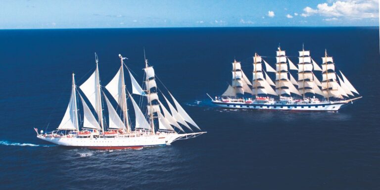 This image features two majestic Star Clippers ships sailing side by side on the open ocean under clear blue skies. Their tall masts are adorned with full white sails, showcasing the elegance of traditional sailing vessels. The scene highlights the beauty and grandeur of cruising aboard these unique, wind-powered ships.