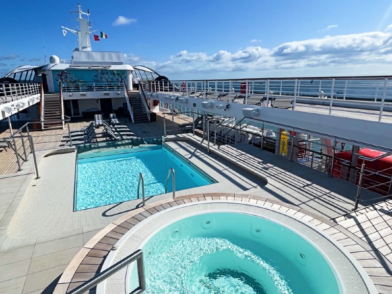 This image captures a clear and inviting view of the pool area on the deck of the Star Legend cruise ship, featuring both a large rectangular swimming pool and a smaller circular hot tub. The deck is laid out with textured tiles that enhance the safety and aesthetics of the space. Sun loungers and open deck areas provide ample space for relaxation and sunbathing under a vast blue sky. The setting is designed for optimal leisure, with the ship's structure and the endless ocean horizon providing a dramatic and tranquil backdrop.