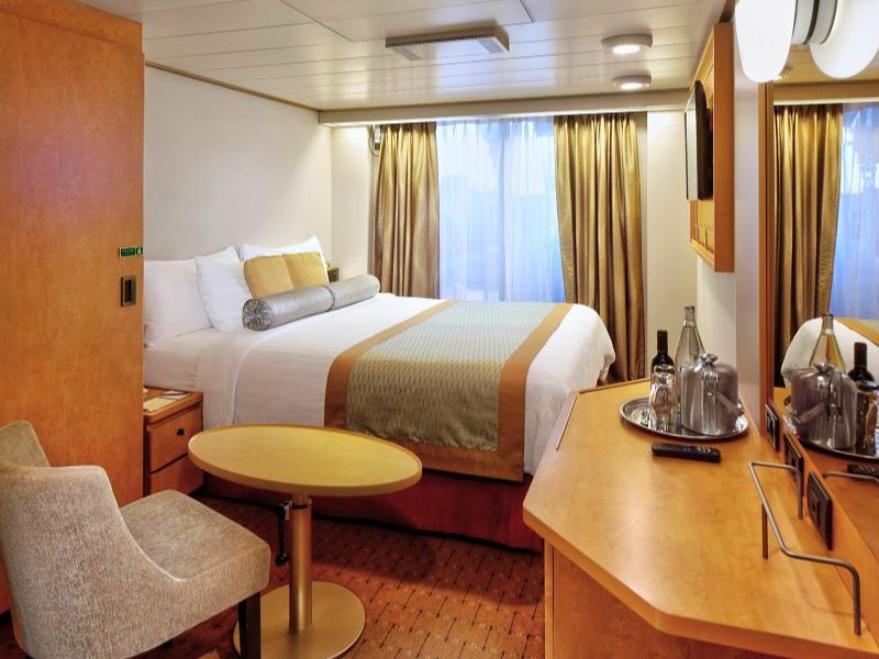 A cosy stateroom aboard a Holland America Line cruise ship, equipped with a comfortable queen-size bed with white and gold bedding, a small seating area with a table and chair, and a vanity mirror with beverage setup.