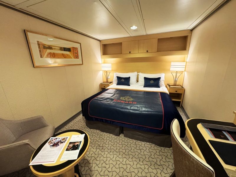 A Cunard cruise ship stateroom featuring a neatly made bed with navy blue bedding and Cunard branding, flanked by two nightstands with lamps, a comfortable chair, and a table with cruise brochures.