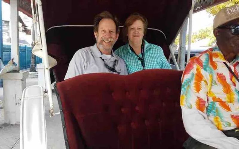Bruce Campbell and his wife smiling while seated in a plush, horse-drawn carriage, with the driver wearing a colorful tropical shirt, capturing a joyful moment of their vacation.