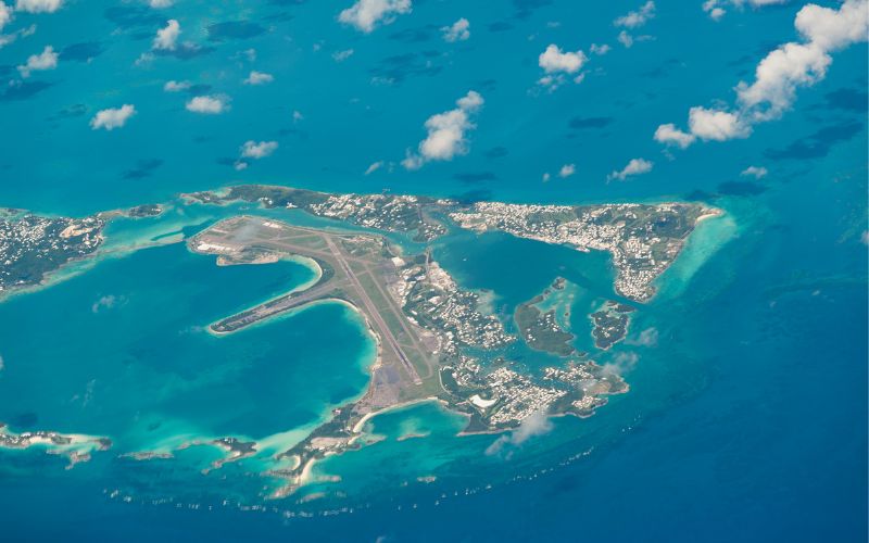 Aerial view of a tropical island in the Bahamas with a clear view of the airport runway, surrounding turquoise waters, and scattered cloud shadows, capturing the essence of being stranded in a picturesque location.