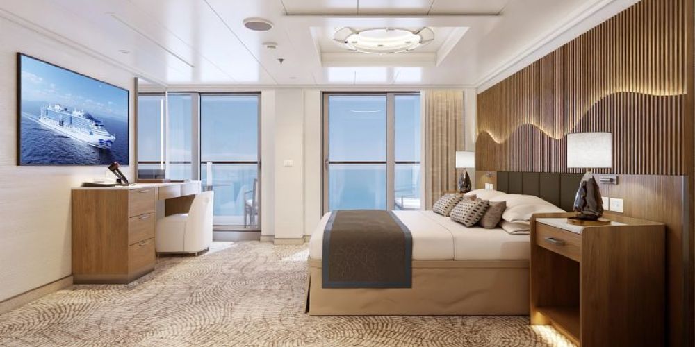 The image shows a luxurious Suite on Princess Cruises' Sun Princess. The suite features a large bed with plush linens and decorative pillows, set against a stylish wood-paneled accent wall. The room is spacious and includes a desk area with a chair, a large flat-screen TV mounted on the wall, and floor-to-ceiling glass doors leading to a private balcony with outdoor seating. The decor is elegant, with a combination of warm wood tones and neutral colors, creating a sophisticated and serene environment. The suite is well-lit, with modern lighting fixtures adding to the overall ambiance of comfort and luxury.