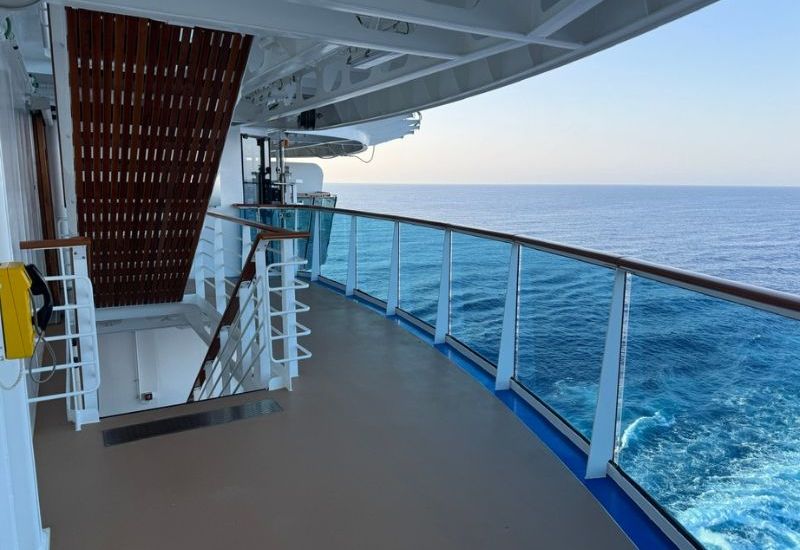 A view from the deck of the Sun Princess cruise ship shows a walkway with glass railings overlooking the ocean. The deck is spacious and clean, with stairs leading to another level. The sea is calm, and the vast expanse of water stretches out to the horizon, creating a peaceful and serene atmosphere aboard the ship.